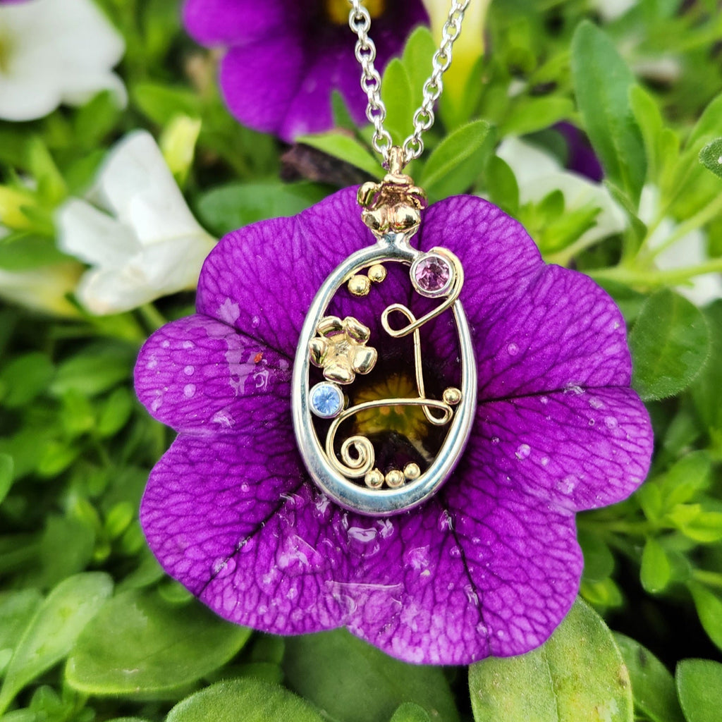 On a purple flower 0utside. A Sterling Silver Oval Frame Pendant with a 14 Karat Yellow Gold Bryn's Blossom flower bale. Interrupting the frame on the top left is a bezel set Pink Tourmaline. Caressing the Tourmaline and making two loops across the frame to swirl at the bottom is an 14 Karat Yellow Gold Wire Swirl. Another 14K Yellow Gold Bryn's Blossom is set in the middle, accompanied by a Ceylon Sapphire. Yellow Gold Beading accenting the inside of the frame. Made by our Craftsman Sydney.