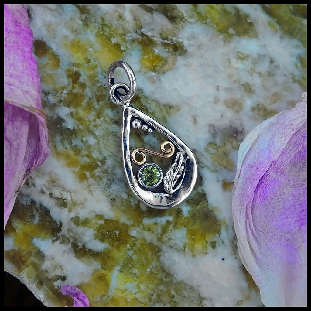 Upon Peony Petals and a slab of Connemara Marble, a small Tear Drop Frame in Sterling Silver. In the lower left is a Sterling Silver Feather charm, adjacent to the feather is a bezel set Peridot. Immediately above lies an 18 Karat Yellow Gold Spiral. Lastly there are three descending Sterling Silver beads on the inner top left of the frame. This pendant includes an 18" Cable Chain. Made by Sydney.