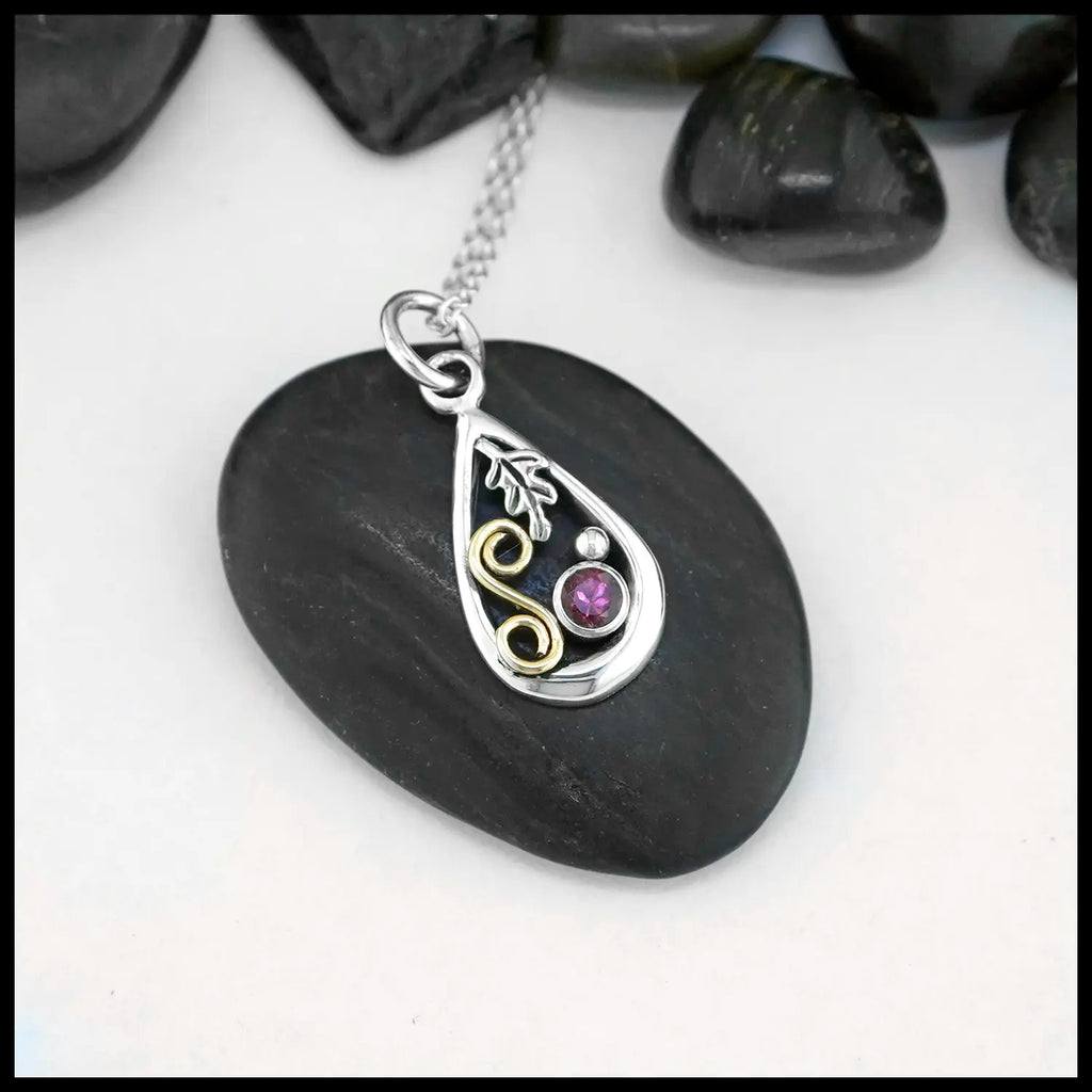 A small Tear Drop Frame in Sterling Silver. In the upper left is a Sterling Silver Oak leaf charm, below the Oak Leaf is a bezel set pink Tourmaline and a Sterling Silver beads. Filling the right side of the frame is an 18 Karat Yellow Gold Spiral. This pendant includes an 18" Cable Chain. Made by Sydney.