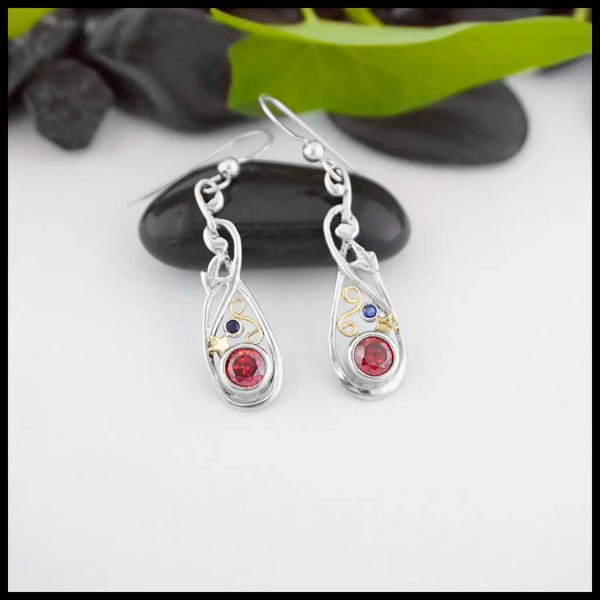 Sterling Silver Nouveau Fleur Frame Drop Earrings with a 5mm Synthetic Garnet and a 2mm Synthetic Sapphire. combining the two stones in the center are a 18K Yellow Gold Swirl and Star. Giving it that Fire Cracker Flare. Fresh off of Terry's Bench.
